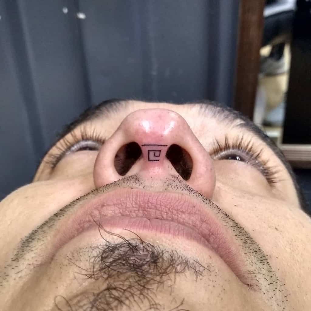 Man's nose tattoo featuring a small geometric square pattern with lines, positioned between the nostrils.
