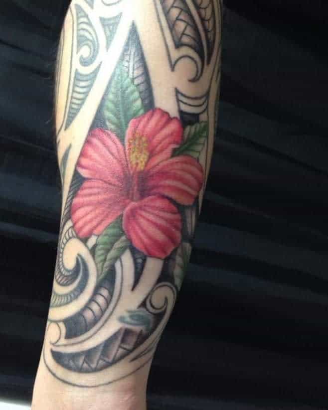 Colorful hibiscus tattoo with tribal patterns on an arm, featuring green leaves and intricate black designs
