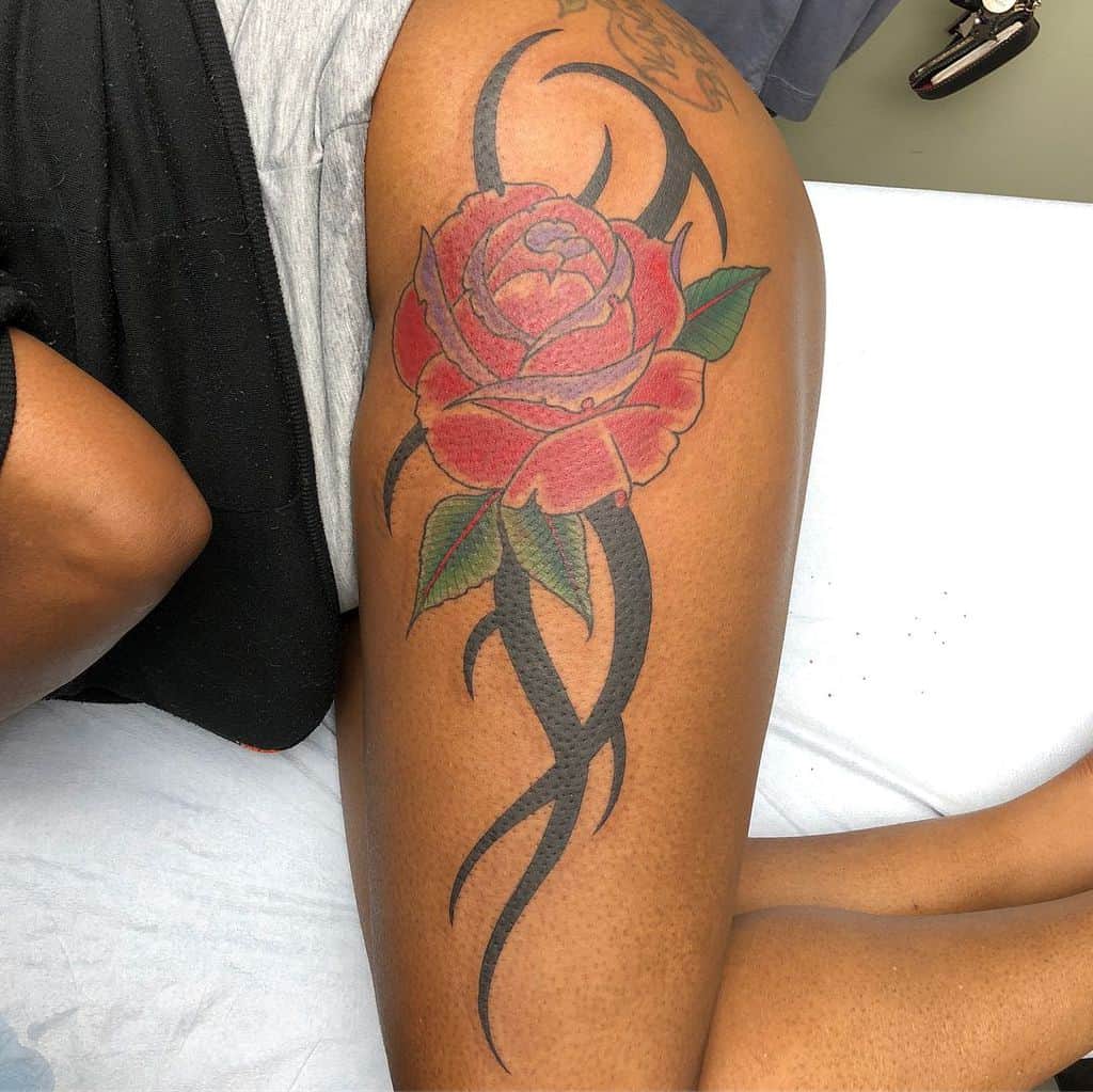 Tattoo of a red rose with green leaves and black tribal patterns on a woman's upper thigh
