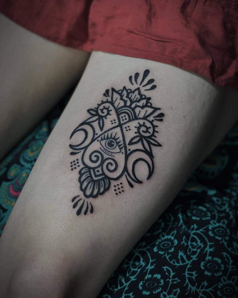 Black intricate geometric tattoo with an eye design on a woman's thigh, surrounded by floral patterns
