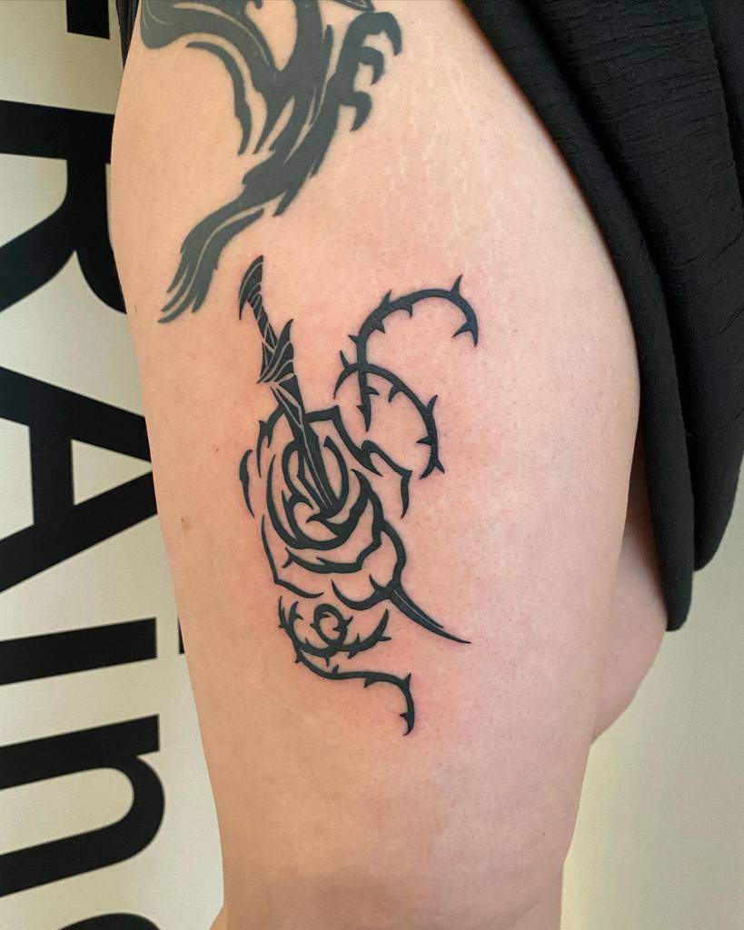 Black-and-gray tattoo of a rose with a dagger through it on an upper arm, surrounded by thorny vines