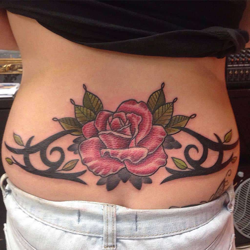 A colorful tattoo of a red rose with green leaves and black tribal patterns on a lower back