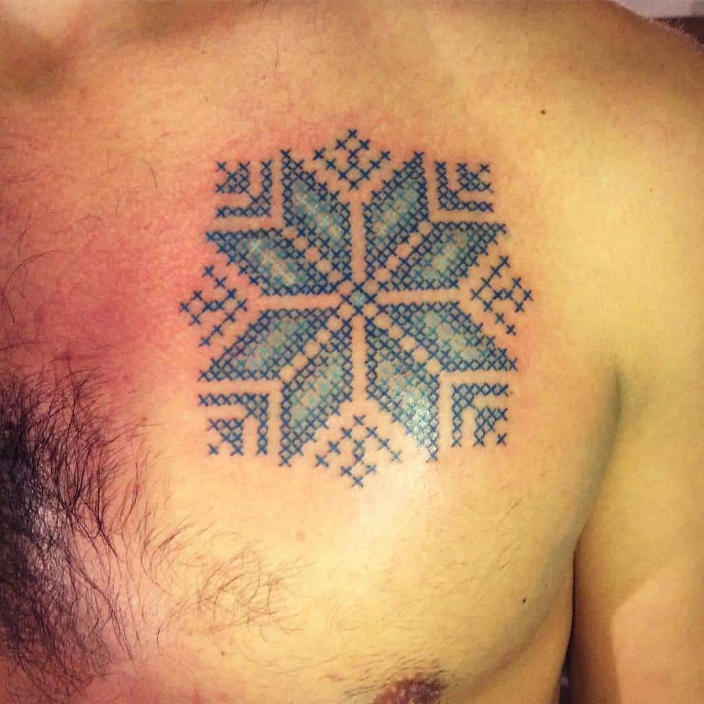 Cross-stitch tattoo of a geometric snowflake pattern in blue, designed to mimic traditional embroidery with intricate detailing