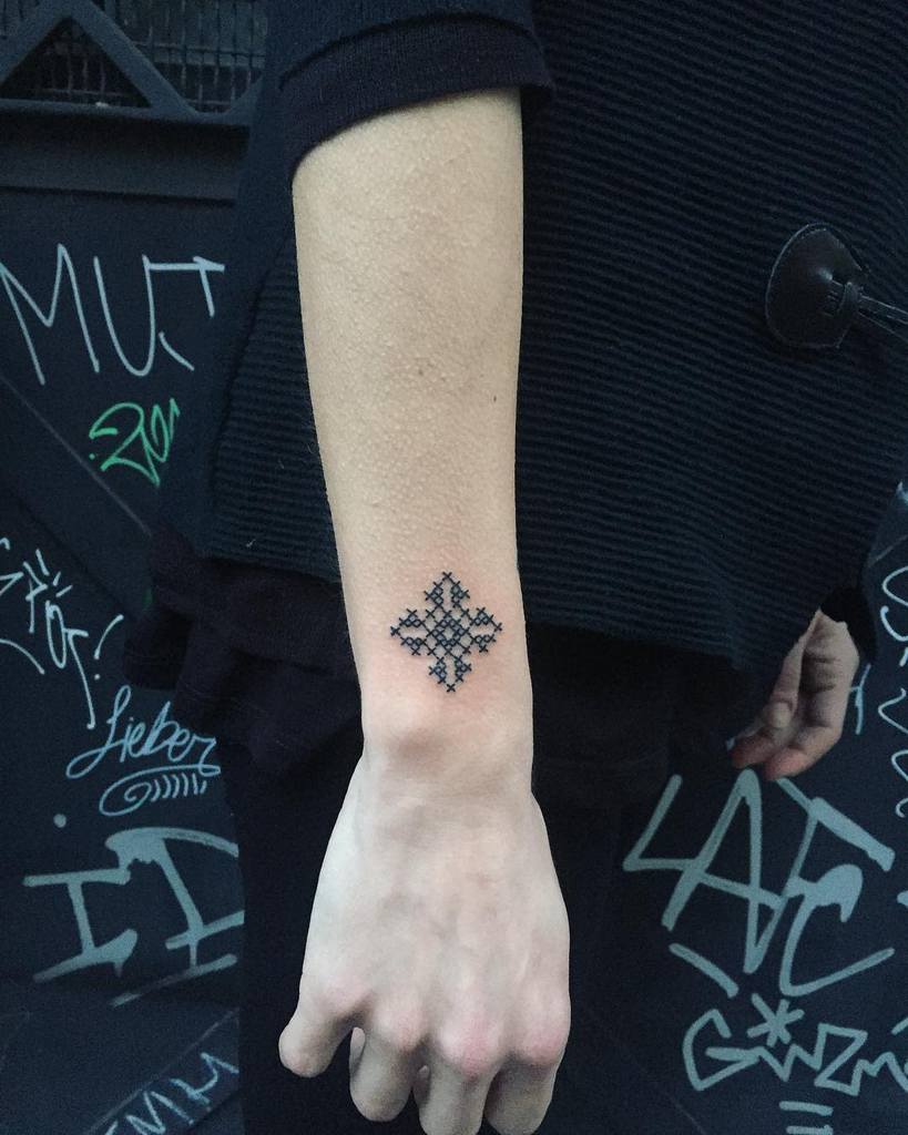 Minimalist cross-stitch tattoo of a small black geometric pattern, designed to resemble traditional embroidery stitches