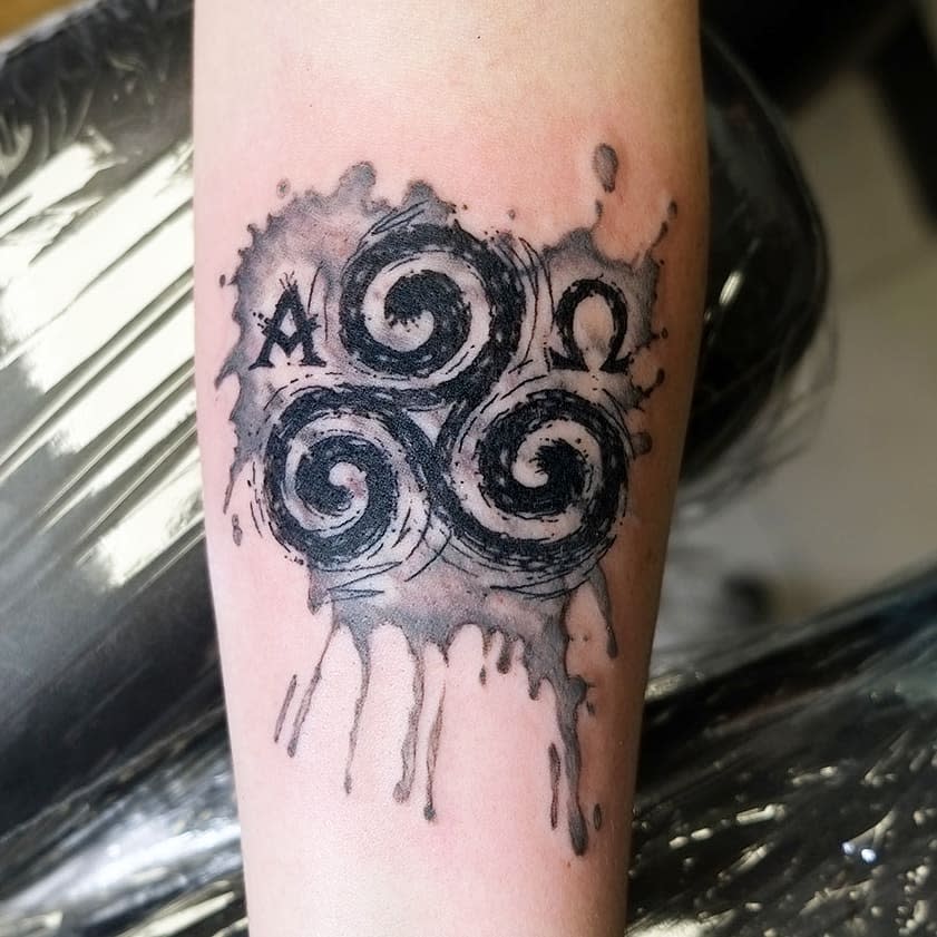 "Forearm tattoo of a Triskelion with watercolor effects, flanked by Alpha and Omega symbols, representing eternal cycles and balance