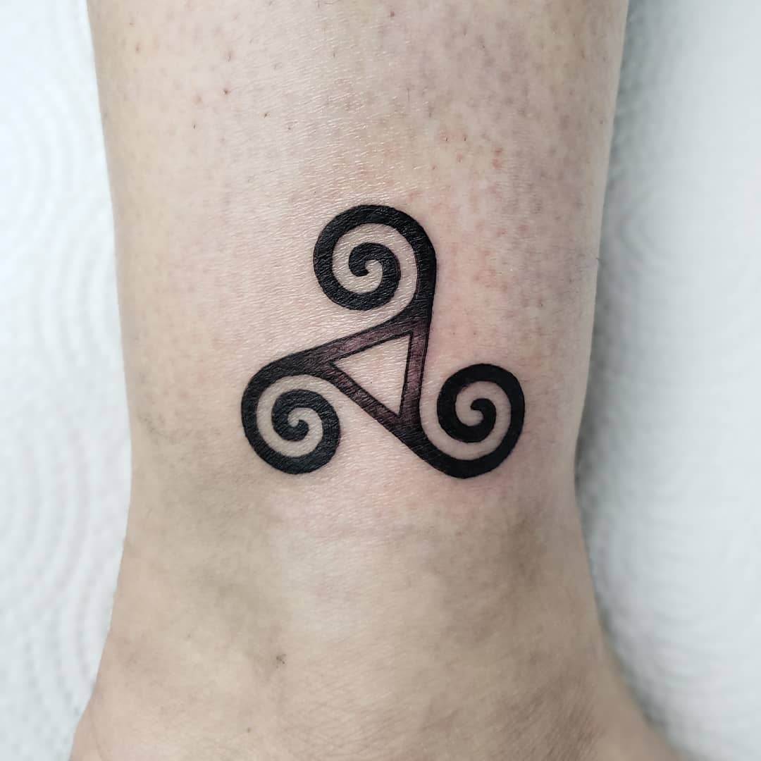 Small tattoo of a black triskelion symbol with spirals on ankle