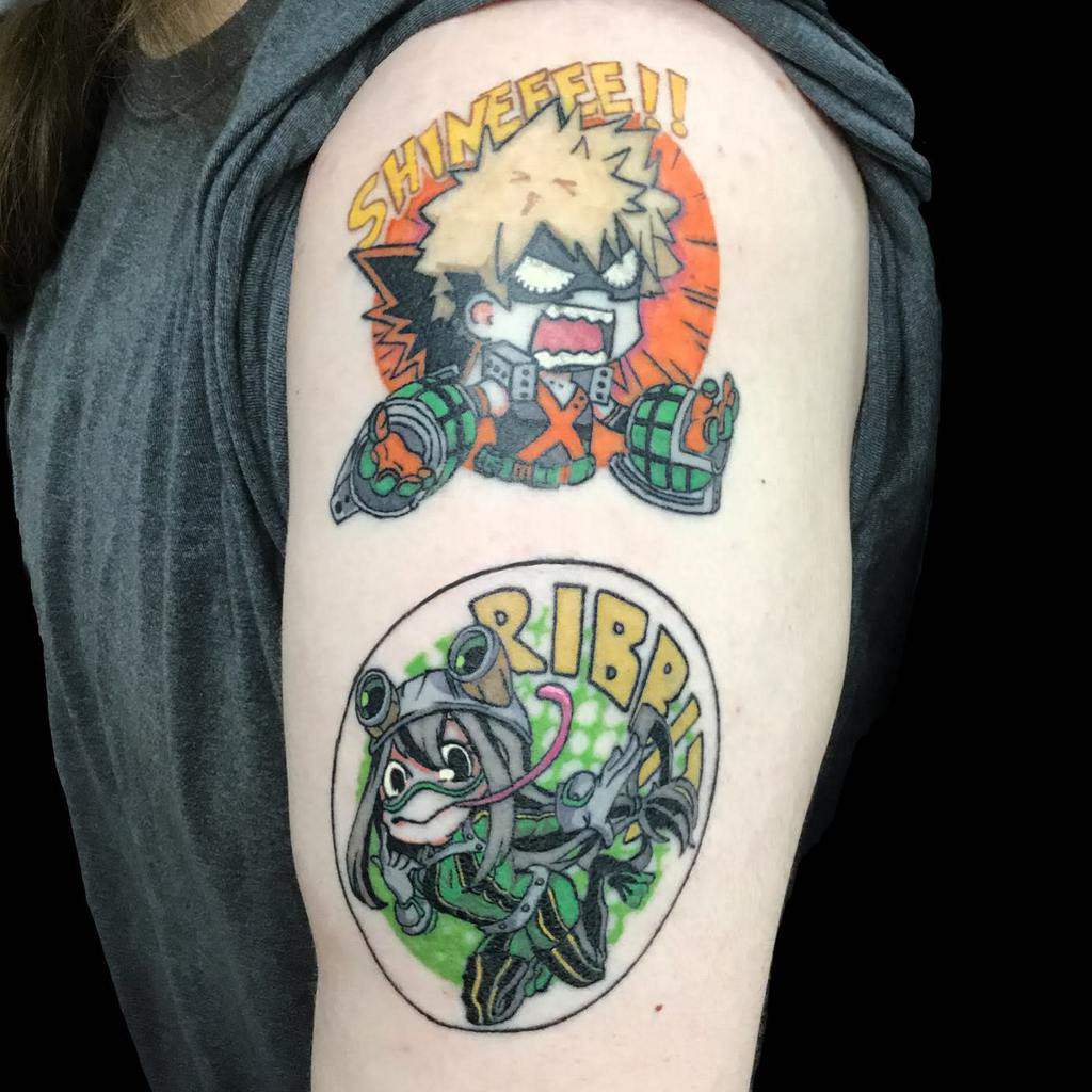 Colorful tattoo featuring chibi-style Katsuki Bakugo shouting 'SHINEEEE!!' with an explosion orange circle background and Tsuyu Asui saying 'RIBBIT' in a frog-like pose with green and white circle background