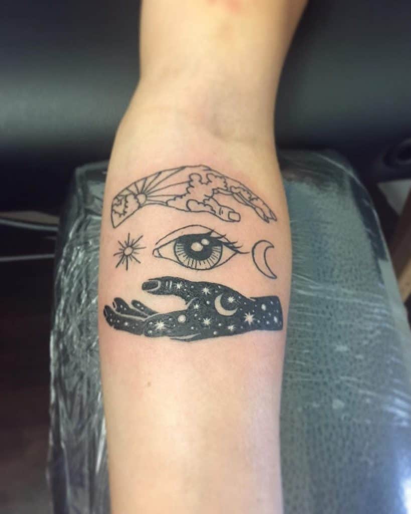 Minimalist black ink tattoo featuring a sun and moon-themed hand, an all-seeing eye, celestial symbols, and a cosmic hand with stars.
