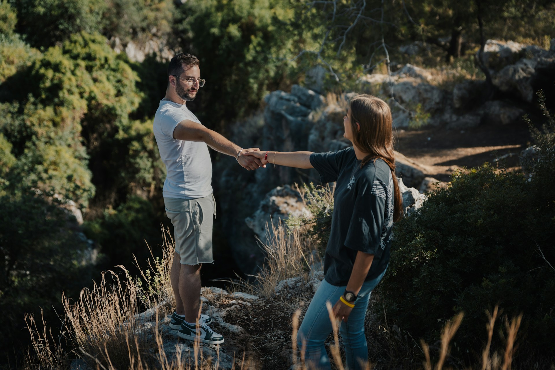 Two people stand on rocky ground, holding hands and facing each other among greenery and sunlight, expressing commitment and support