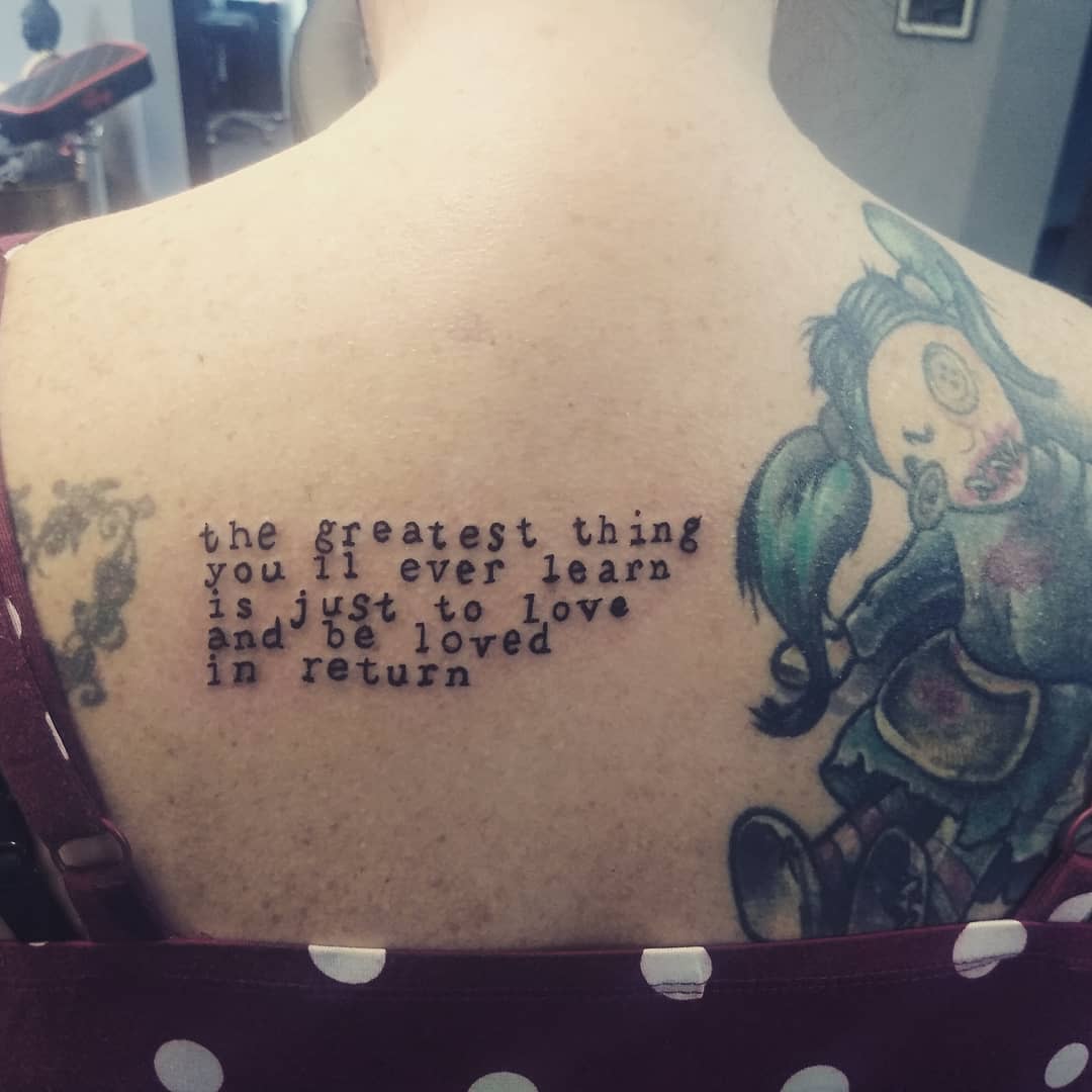 Tattoo on a back with a quote: "the greatest thing you'll ever learn is to love and be loved in return" and a colorful doll illustration