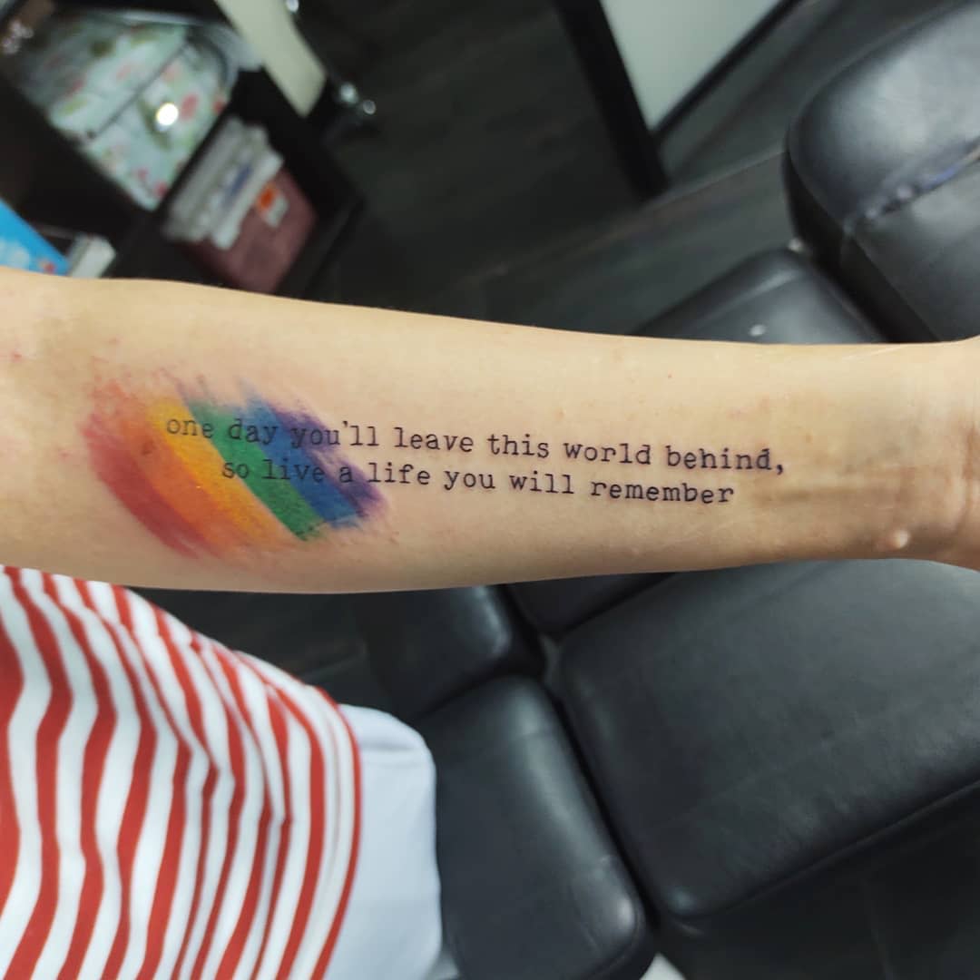 Forearm tattoo featuring rainbow brush strokes and a motivational quote in black text
