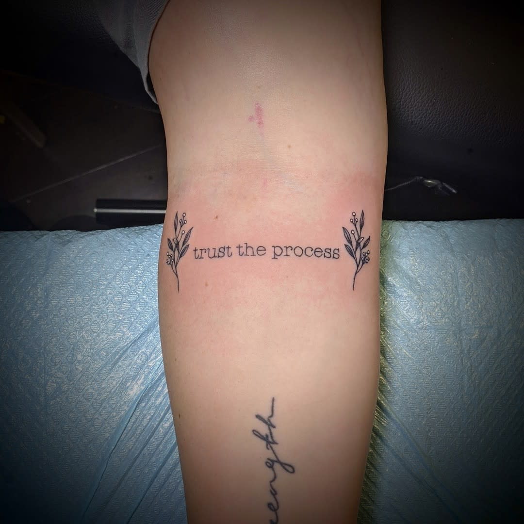A "trust the process" tattoo with small branches on either end on a leg