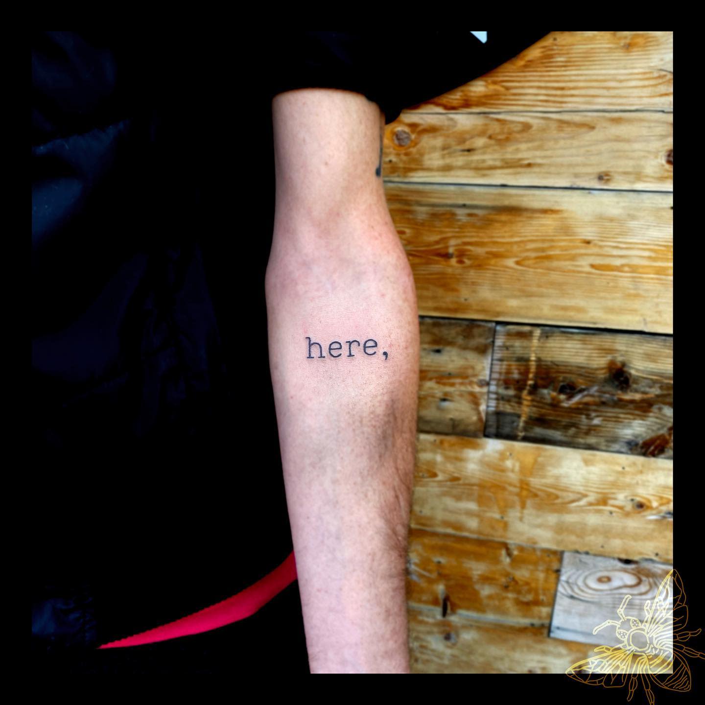 Tattoo of the word "here," on an inner forearm with a wooden background