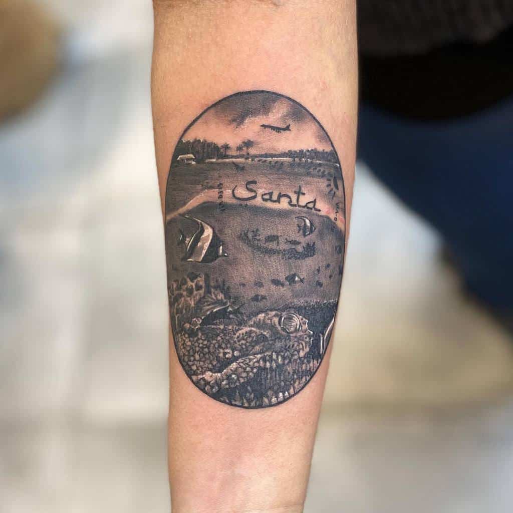 Black and gray oval-shaped tattoo of an ocean and beach scene, featuring fish, a sea turtle, and the word 'Santa' written in the sand
