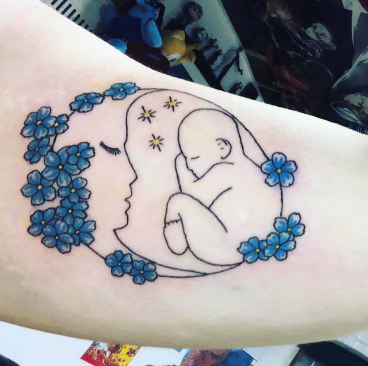 Tattoo of a mother and baby inside a crescent moon surrounded by blue flowers with the mother having a serene profile