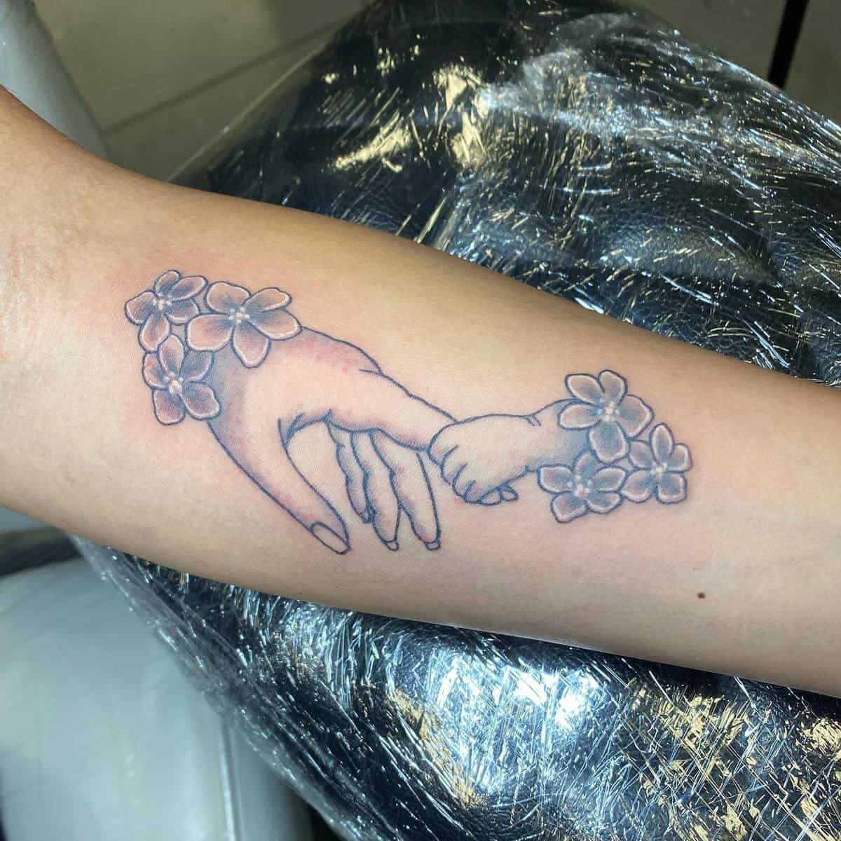 Tattoo of an adult hand and a child's hand holding, decorated with floral designs, on a forearm