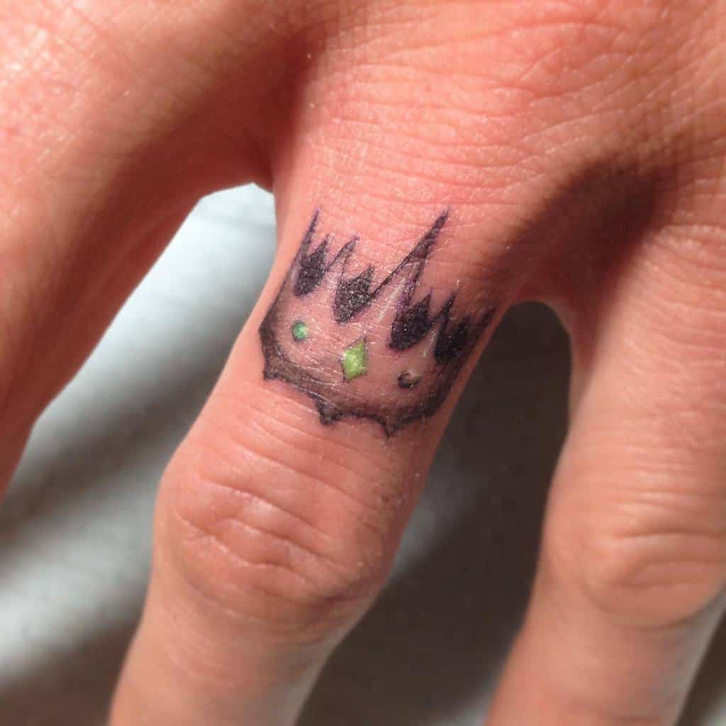 A crown ring tattoo with jagged edges, small colored dots and green diamond in the center