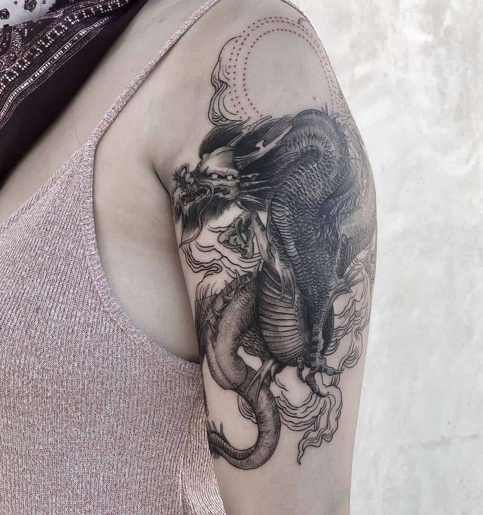 Intricate upper arm tattoo featuring a detailed black and grey dragon surrounded by swirling clouds and fine linework