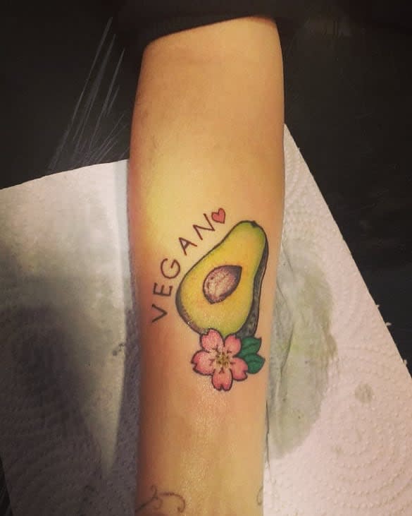 Tattoo of an avocado with a pink flower and the word "VEGAN" on an arm