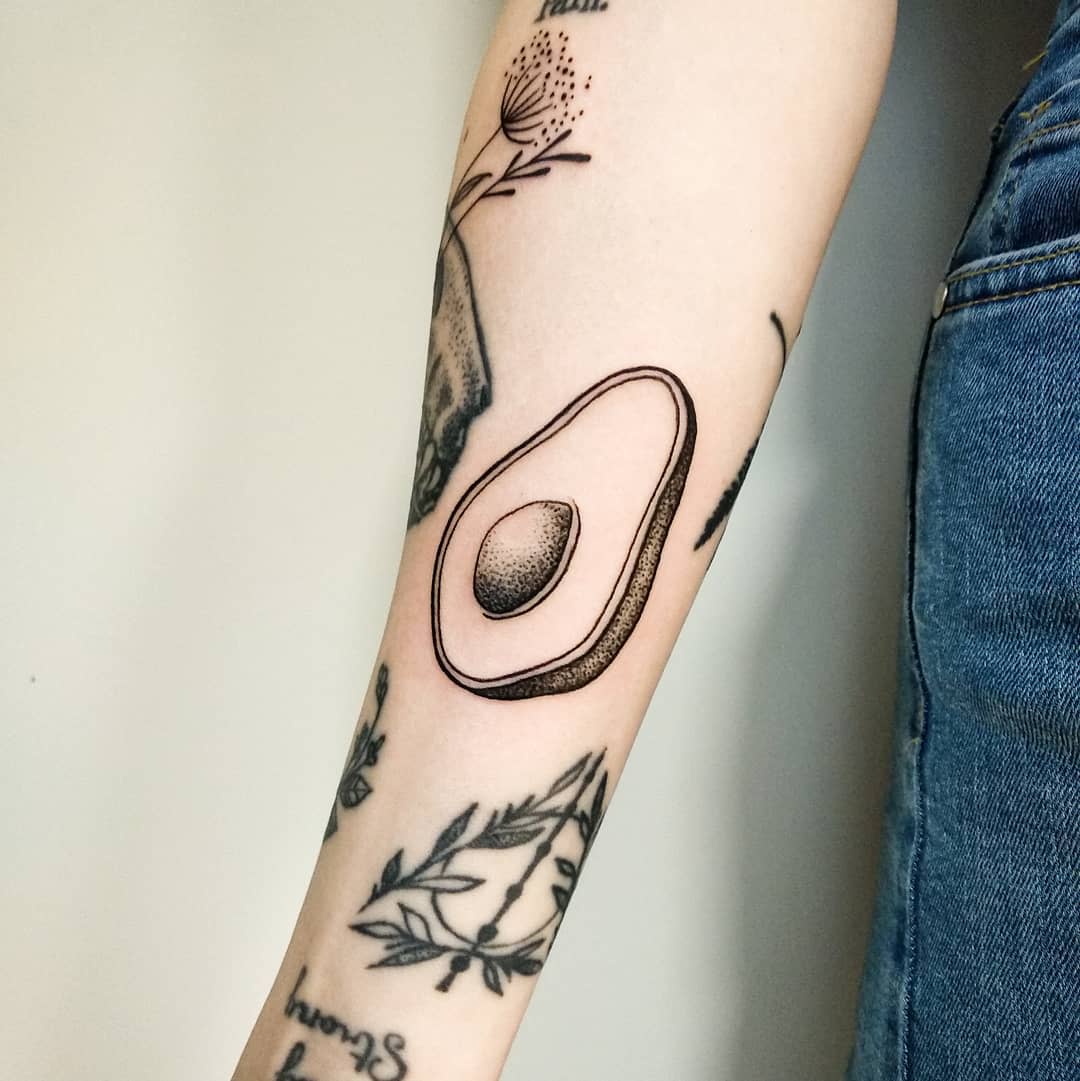Forearm with a detailed avocado tattoo featuring various plant and text tattoos