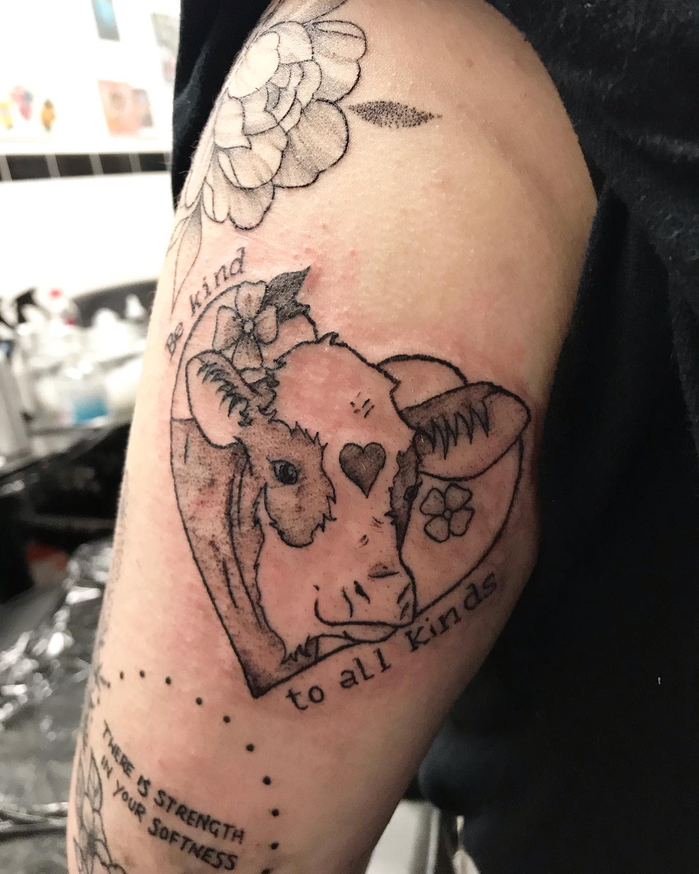 Tattoo of a cow inside a heart with the text "Be kind to all kinds" on an upper arm