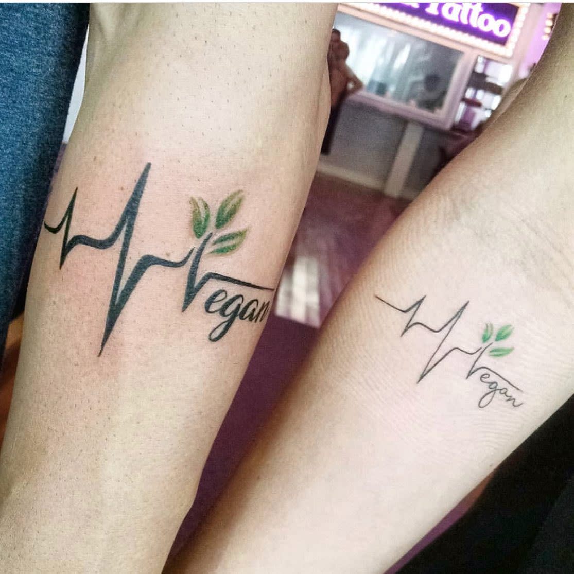 Matching tattoos on two arms with a heartbeat line, leaves, and the word "Vegan"