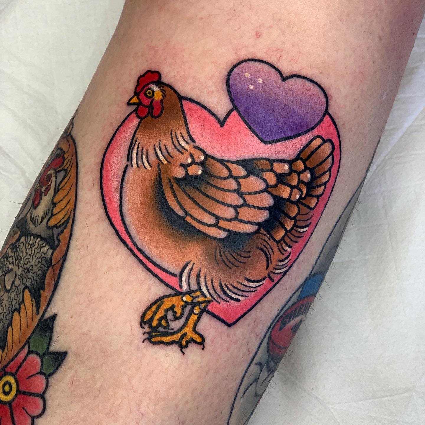 Tattoo of a chicken inside a pink heart with a purple heart above it on an arm