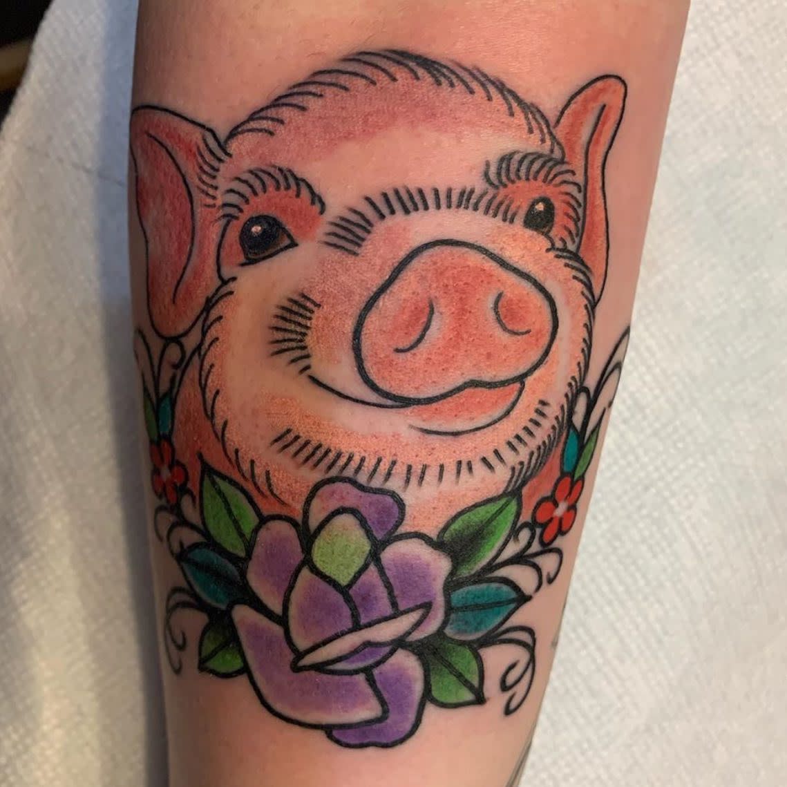 Tattoo of a smiling pig above flowers and leaves on a forearm