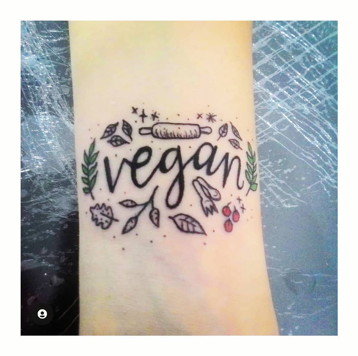 Tattoo on forearm reads "vegan" surrounded by leaves, vegetables, and a rolling pin