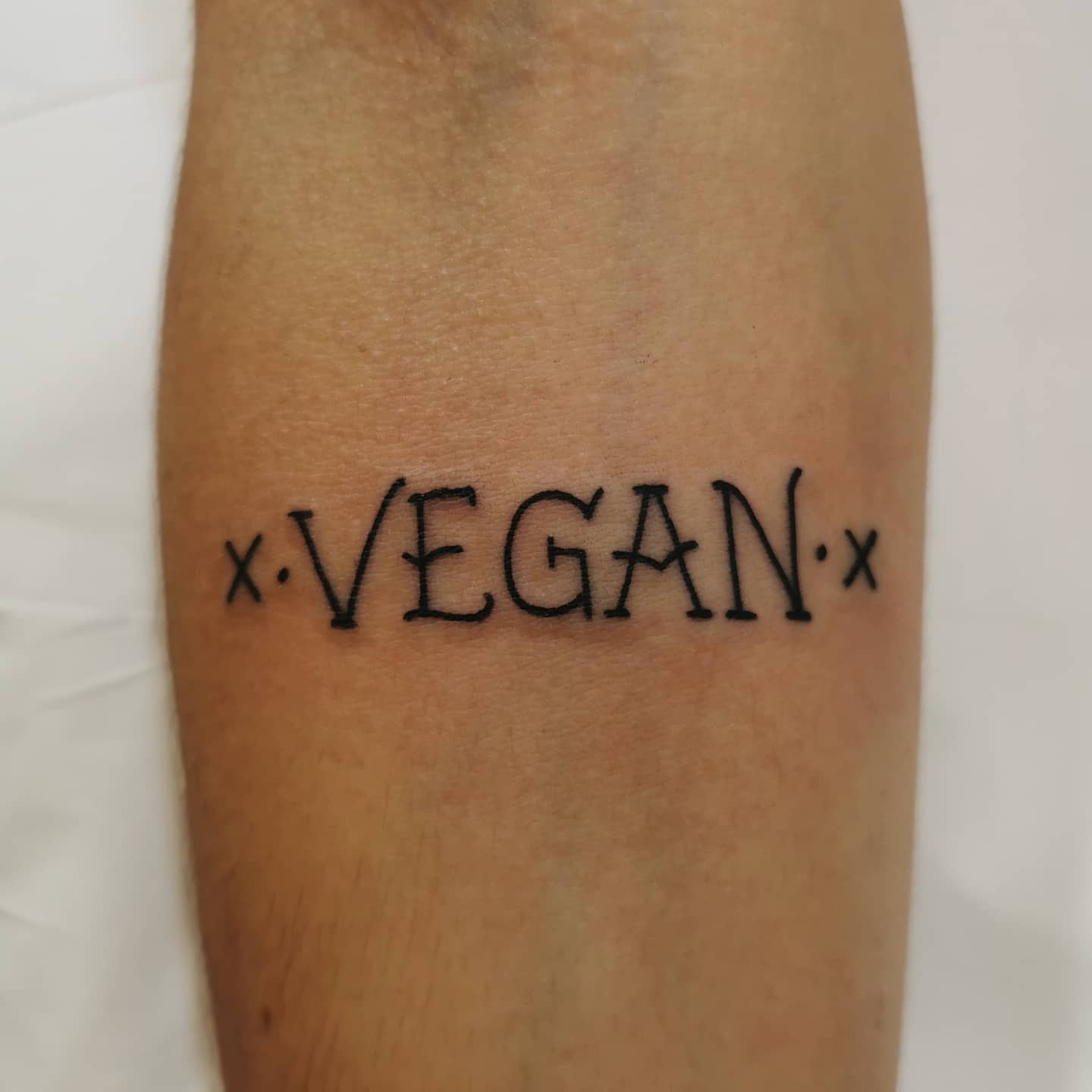 Tattoo on arm reading "VEGAN" with small "x" symbols on either side