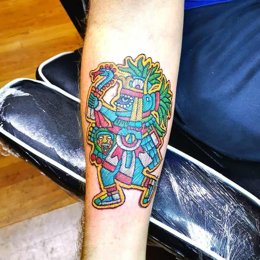 Tattoo of an ancient Mesoamerican deity or warrior, designed with a fabric-like embroidery effect and bold colors.