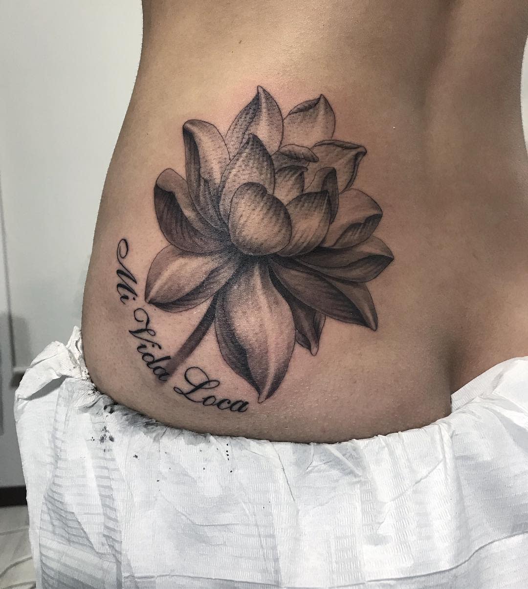 A shaded black and grey water lily tattoo on the hip, accompanied by the text 'Mi Vida Loca' in cursive