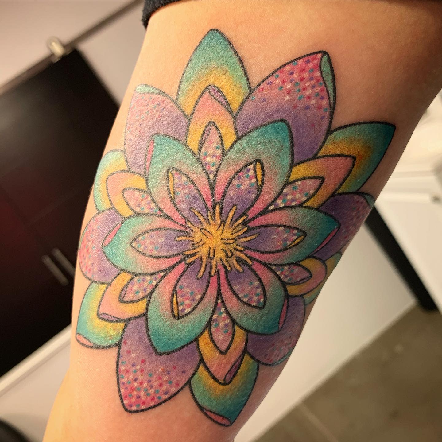 A colorful water lily tattoo on the arm, featuring overlapping petals in shades of teal, pink, purple, and yellow, with intricate dot details and a bright yellow center