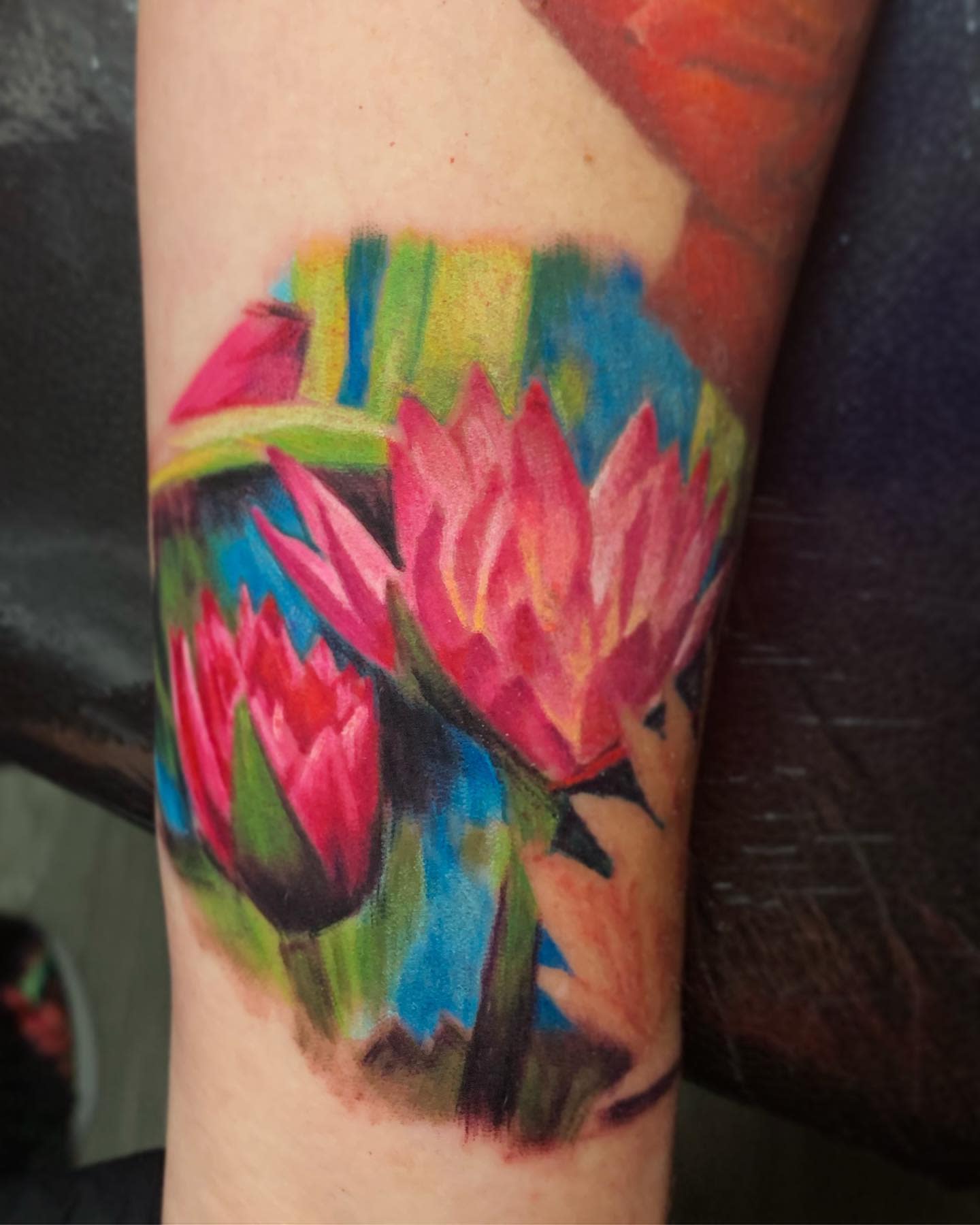 A vibrant and realistic water lily tattoo on the arm, featuring pink blossoms with green leaves and a colorful background of blue and green hues