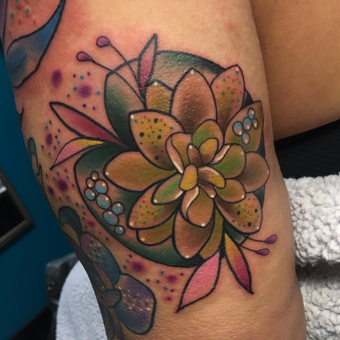 A colorful water lily tattoo on the upper arm, featuring earthy tones of green and brown with pink and blue accents, surrounded by dot details and additional floral elements