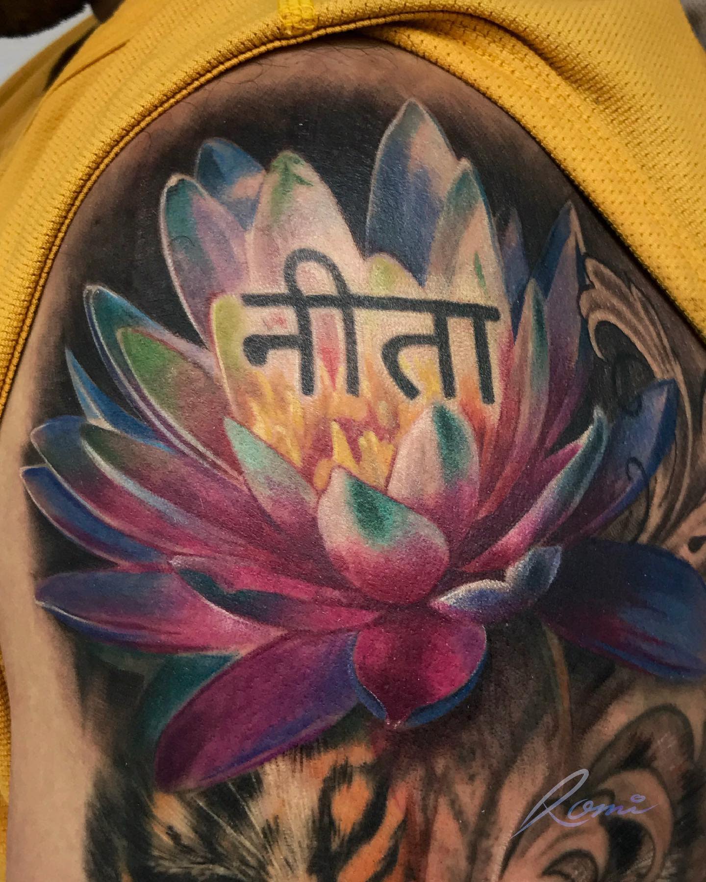A vibrant water lily tattoo on the shoulder, featuring multi-colored petals in shades of blue, purple, pink, and green, with Hindi script above the flower