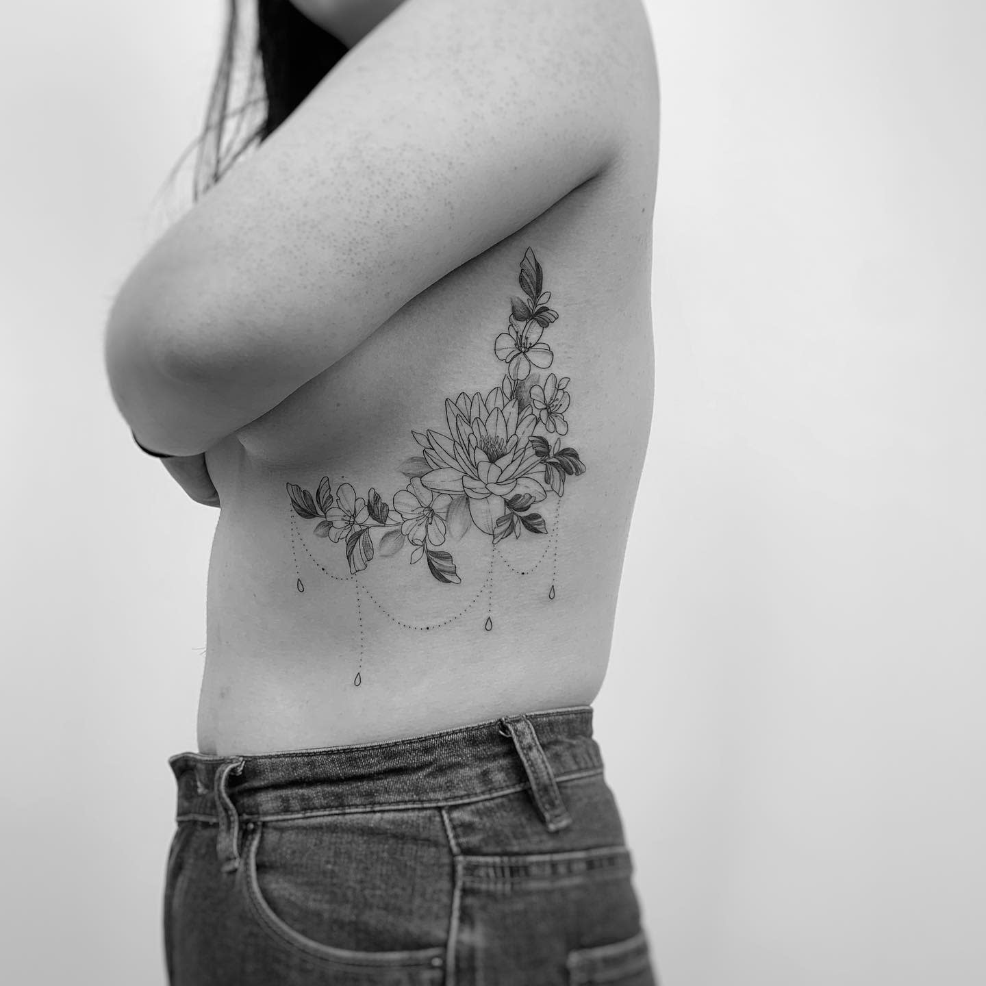 A delicate black and grey water lily tattoo on the ribcage, featuring floral elements, leafy accents, and dotted chains with teardrop details, creating an elegant and feminine design