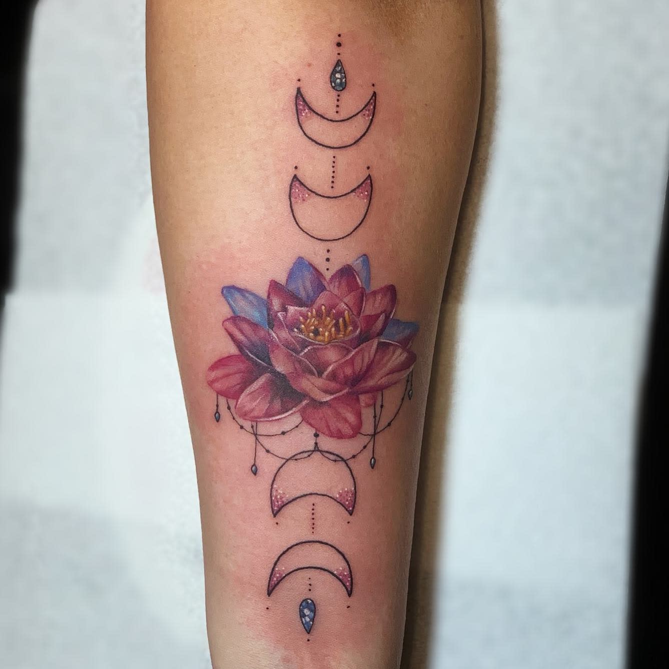 A colorful water lily tattoo on the forearm, featuring red and blue petals, with crescent moon phases and dot work arranged vertically above and below the flower