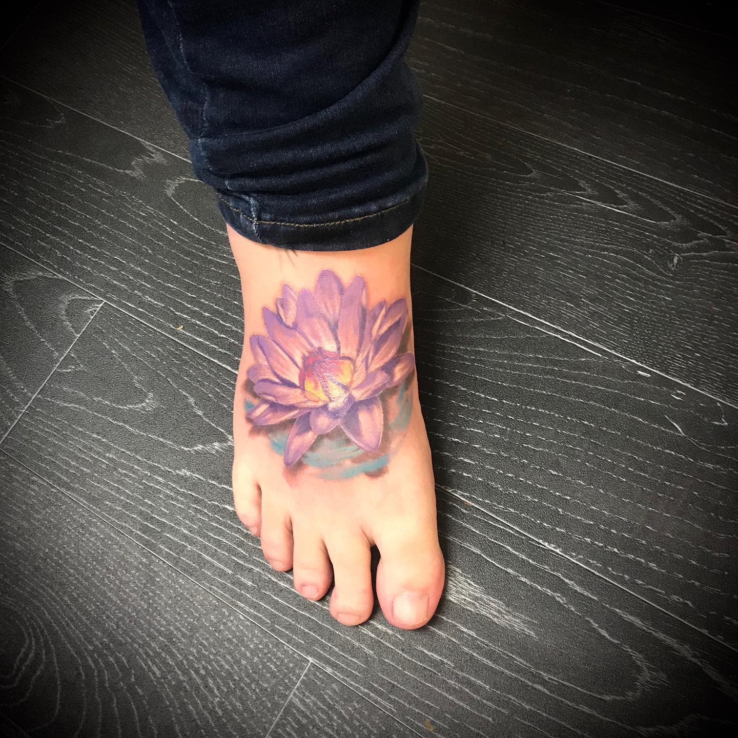 A realistic purple water lily tattoo on the top of the foot, featuring soft shading and a subtle green and blue background