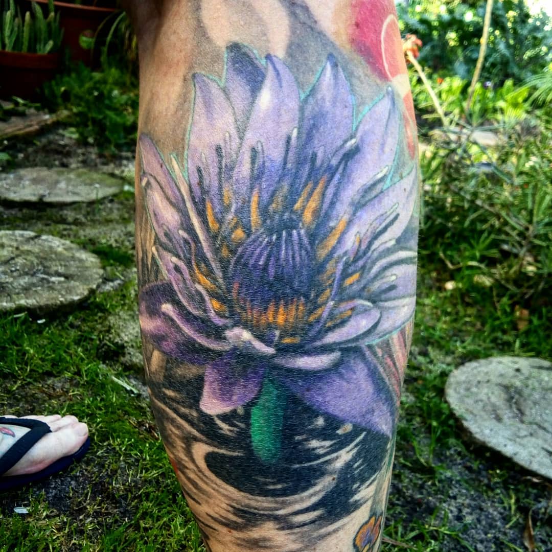 A vibrant purple water lily tattoo on the calf, featuring detailed petals and a dynamic water background with swirling patterns