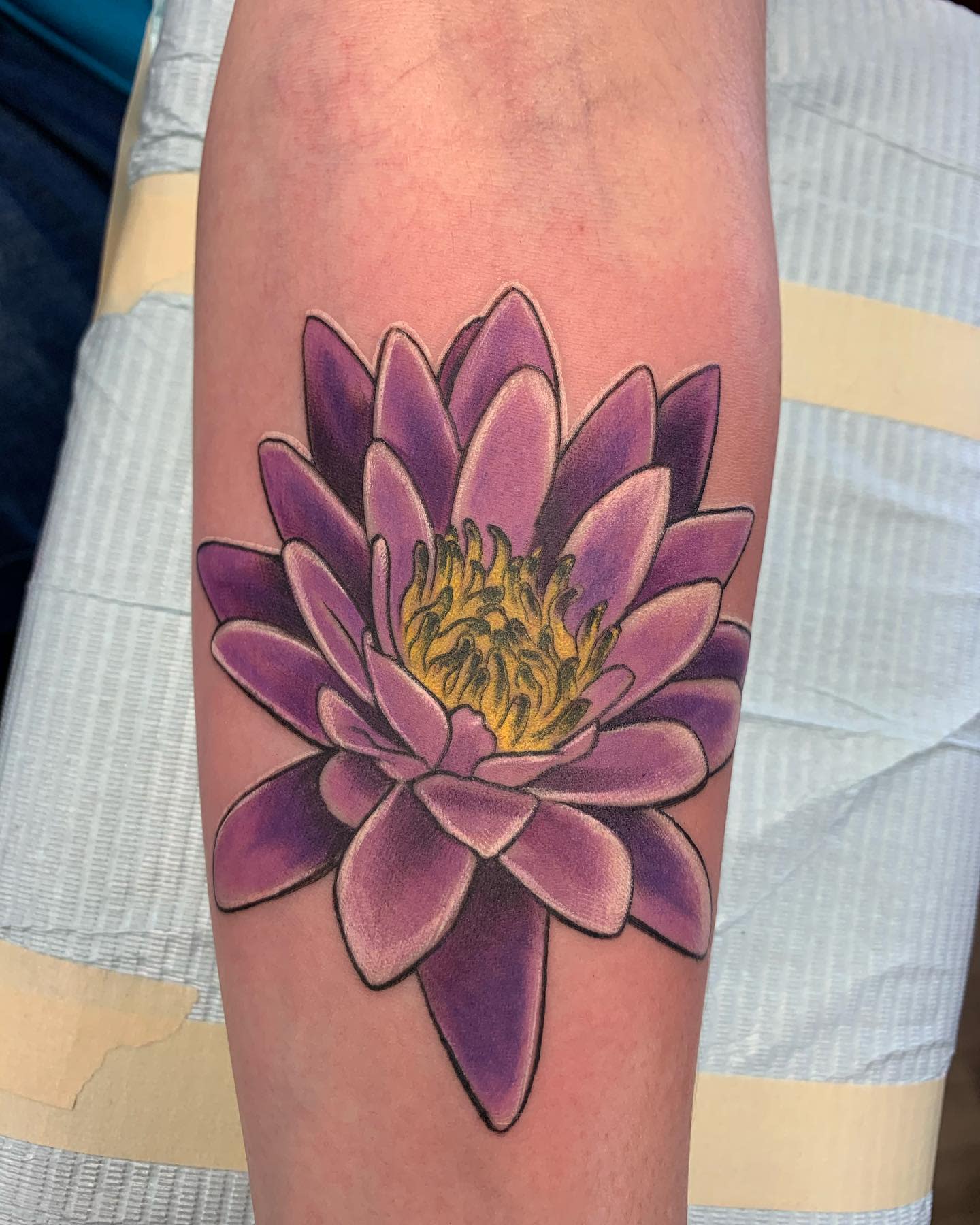 A vibrant purple water lily tattoo on the forearm, featuring bold petals with a bright yellow center and strong black outlines