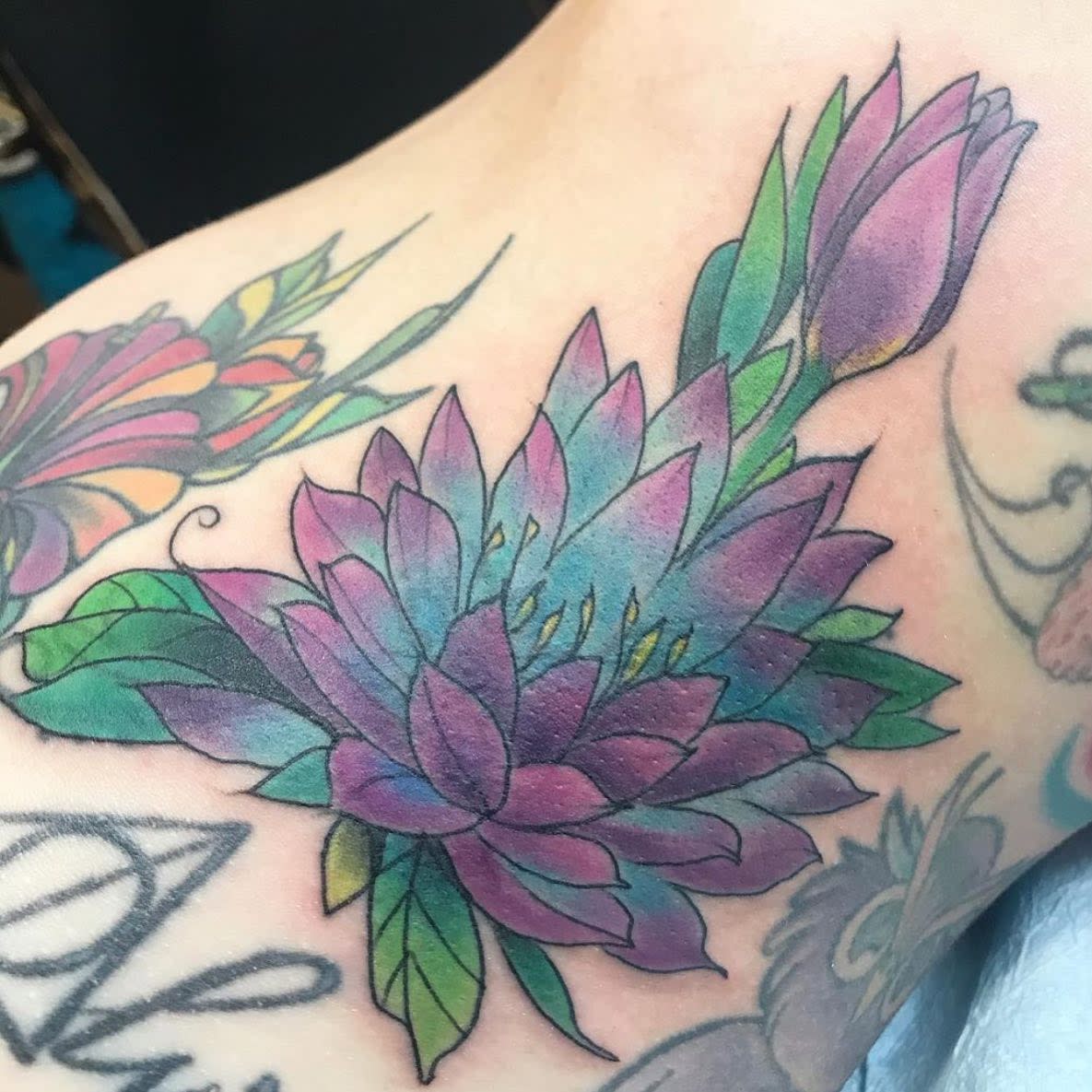 A colorful water lily tattoo on the shoulder, featuring petals in shades of purple, blue, and green with matching leaves and a bud