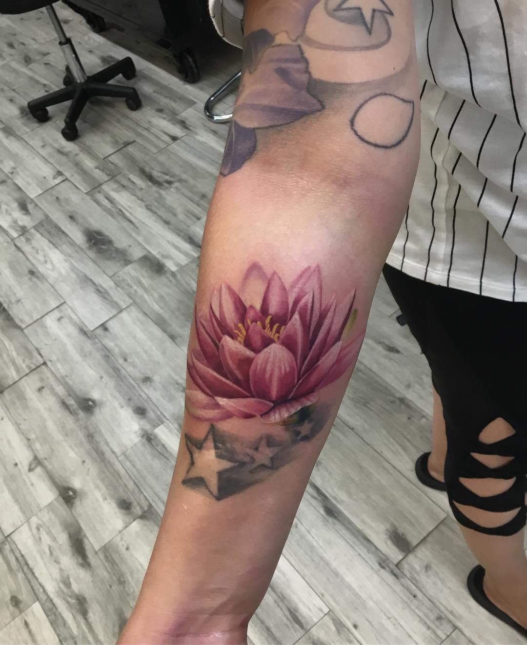 A realistic water lily tattoo on the forearm, featuring deep pink petals and a yellow center, with shaded stars beneath the flower