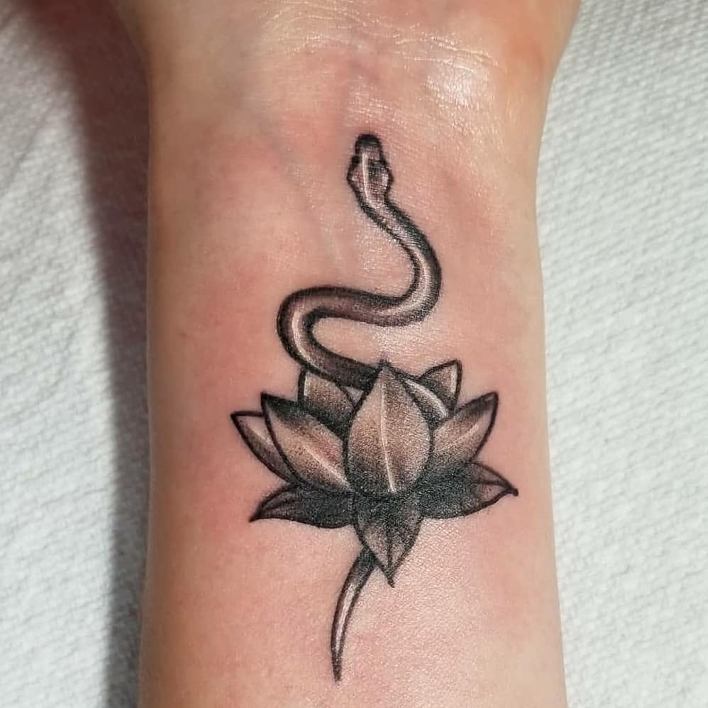 A small black and grey tattoo on the wrist, featuring a water lily with a snake coiled above it, combining elements of nature and symbolism