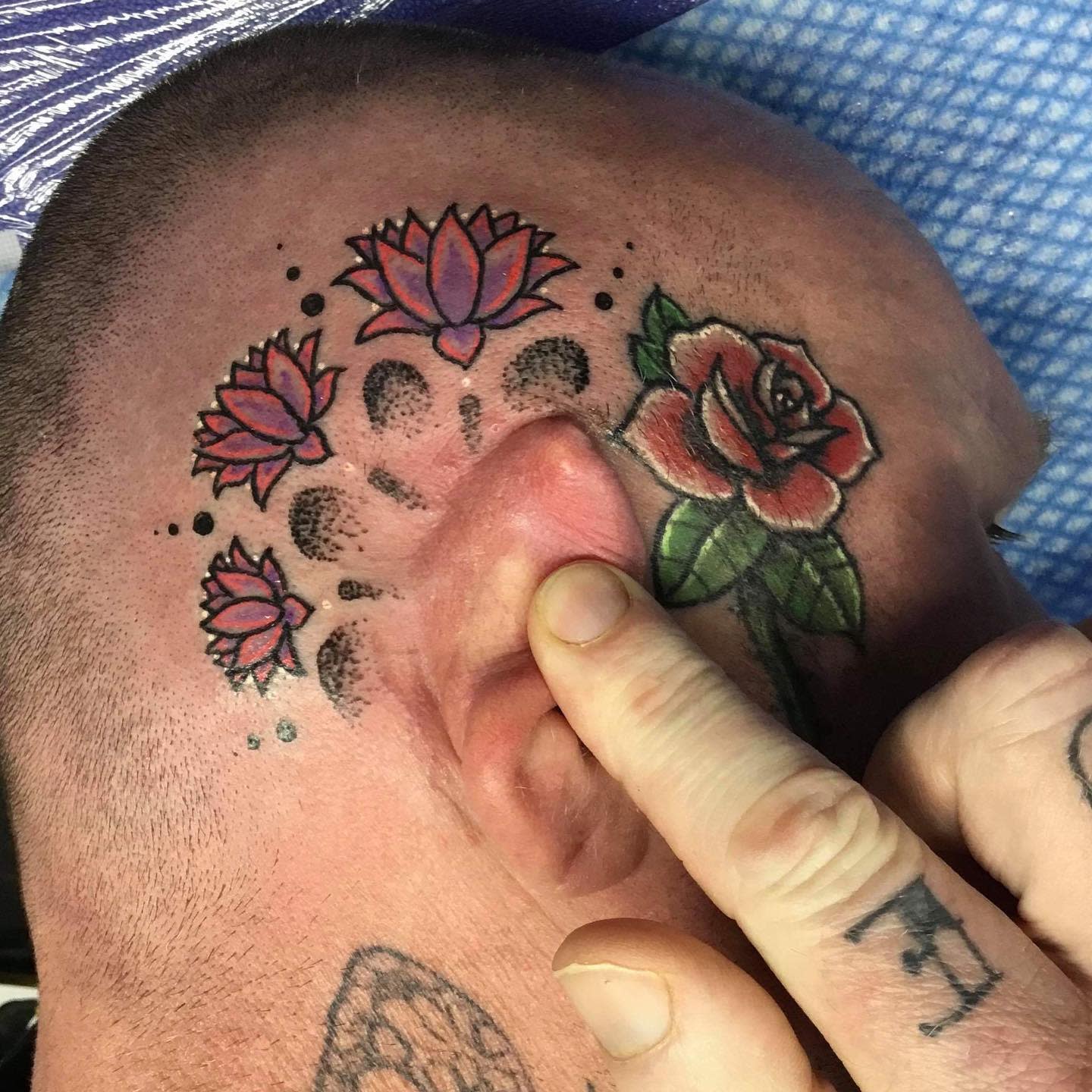 A colorful tattoo on the side of the head, featuring small pink and purple water lilies above the ear, along with a red rose and black dot accents, creating a vibrant and artistic design