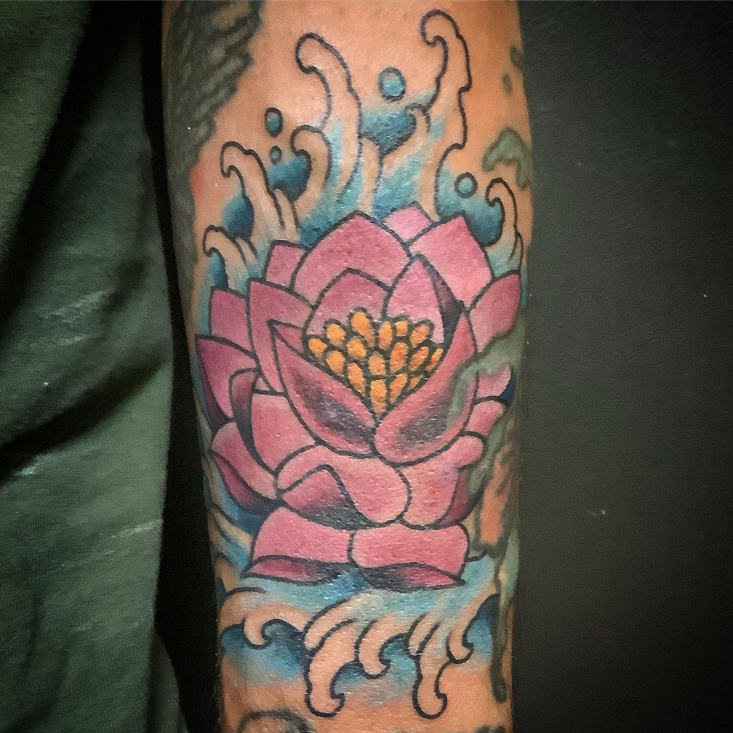 A vibrant traditional-style water lily tattoo on the arm, featuring pink petals, yellow center details, and blue wave accents surrounding the flower