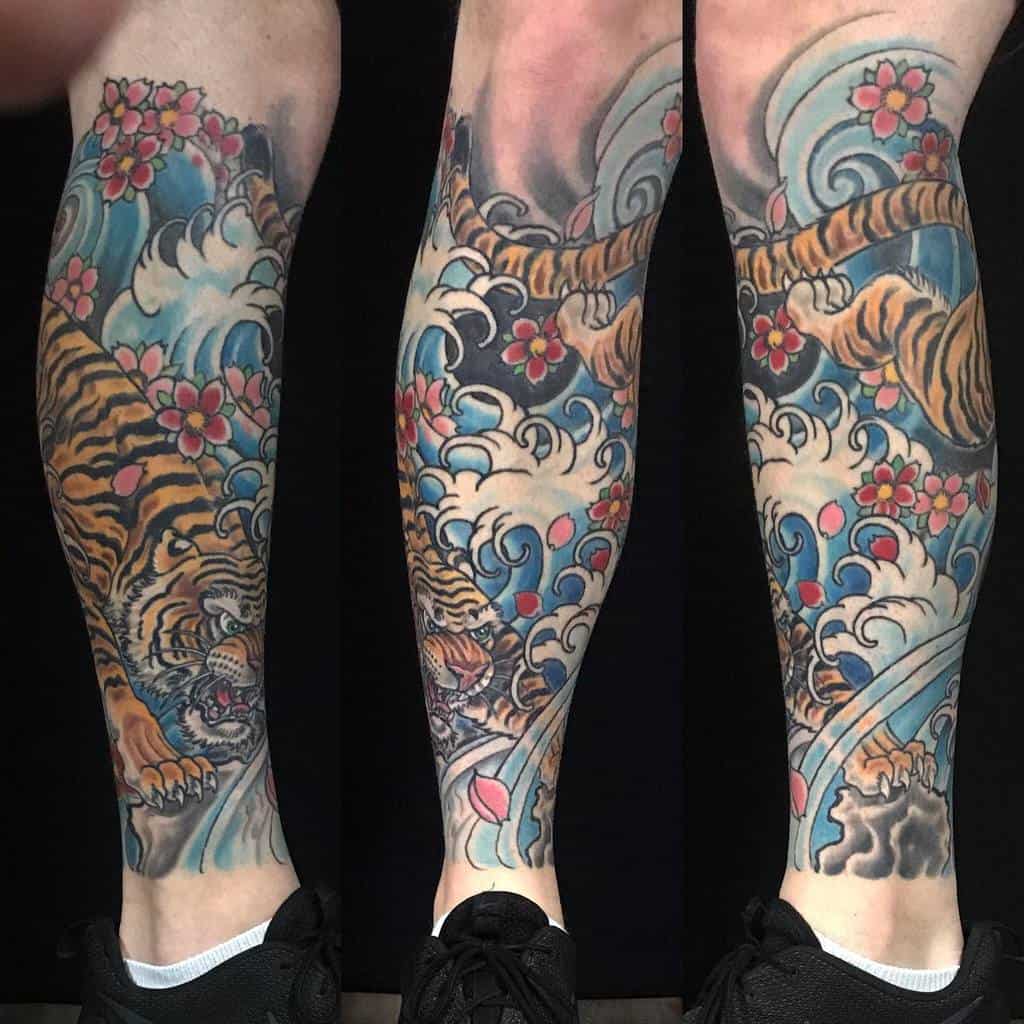 A vibrant Japanese-style leg tattoo featuring a fierce tiger amidst swirling blue water, accented by pink cherry blossoms and dynamic waves