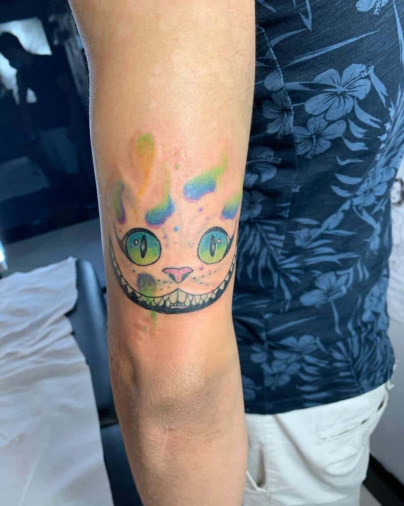Colorful Cheshire Cat tattoo with a wide grin and green eyes on a person's upper arm