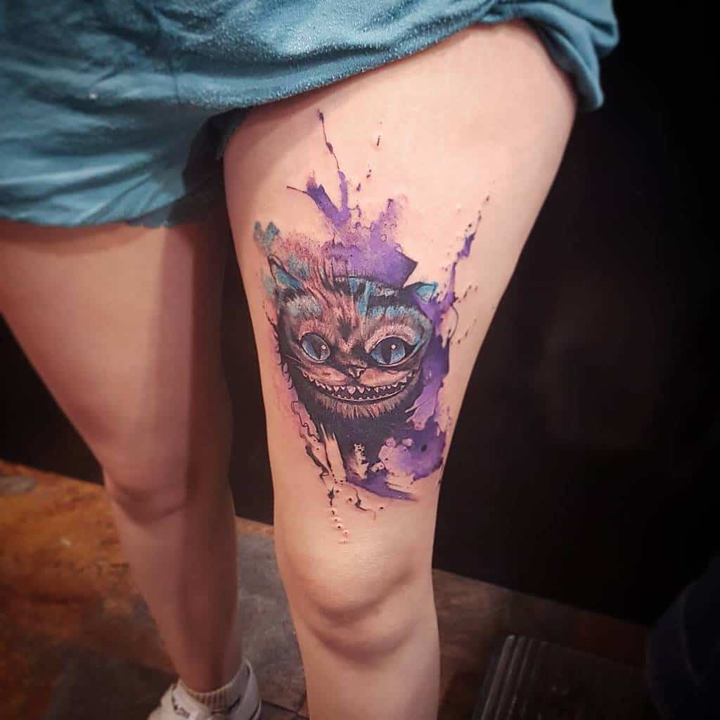 A vibrant watercolor tattoo of the Cheshire Cat on a person's upper thigh