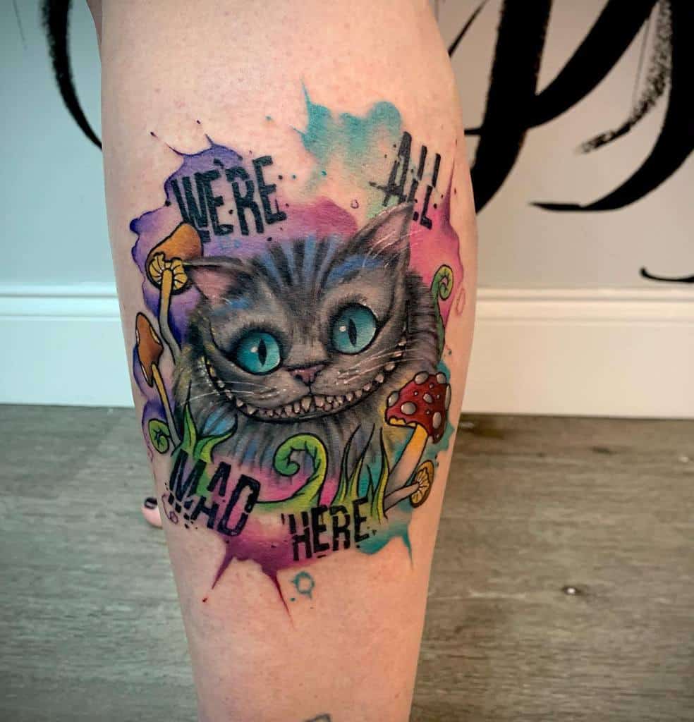 Colorful tattoo of the Cheshire Cat with text "We're all mad here" on a leg